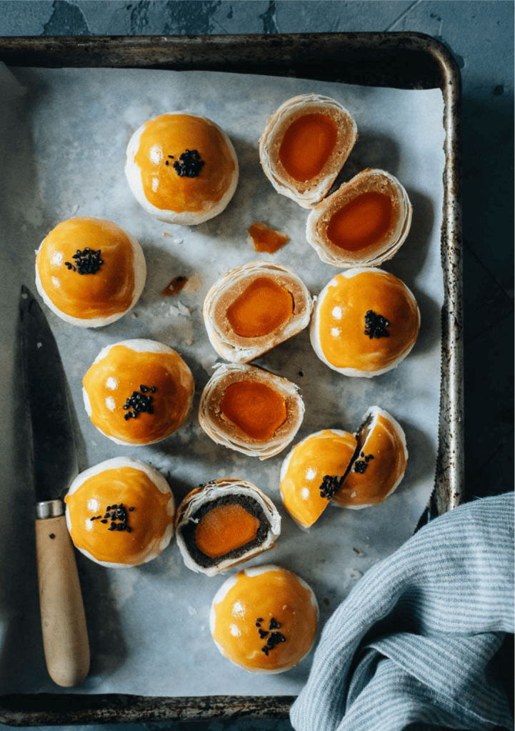 A tray of golden baked pastries with shiny tops, some whole and others cut in half revealing orange fillings. A knife and a blue cloth are also visible.
